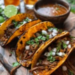 A close-up photo of four delicious birria tacos on a rustic wooden board, topped with fresh cilantro, diced onions, and served with lime wedges. A small bowl of tangy consommé and a pitcher of horchata are placed nearby.